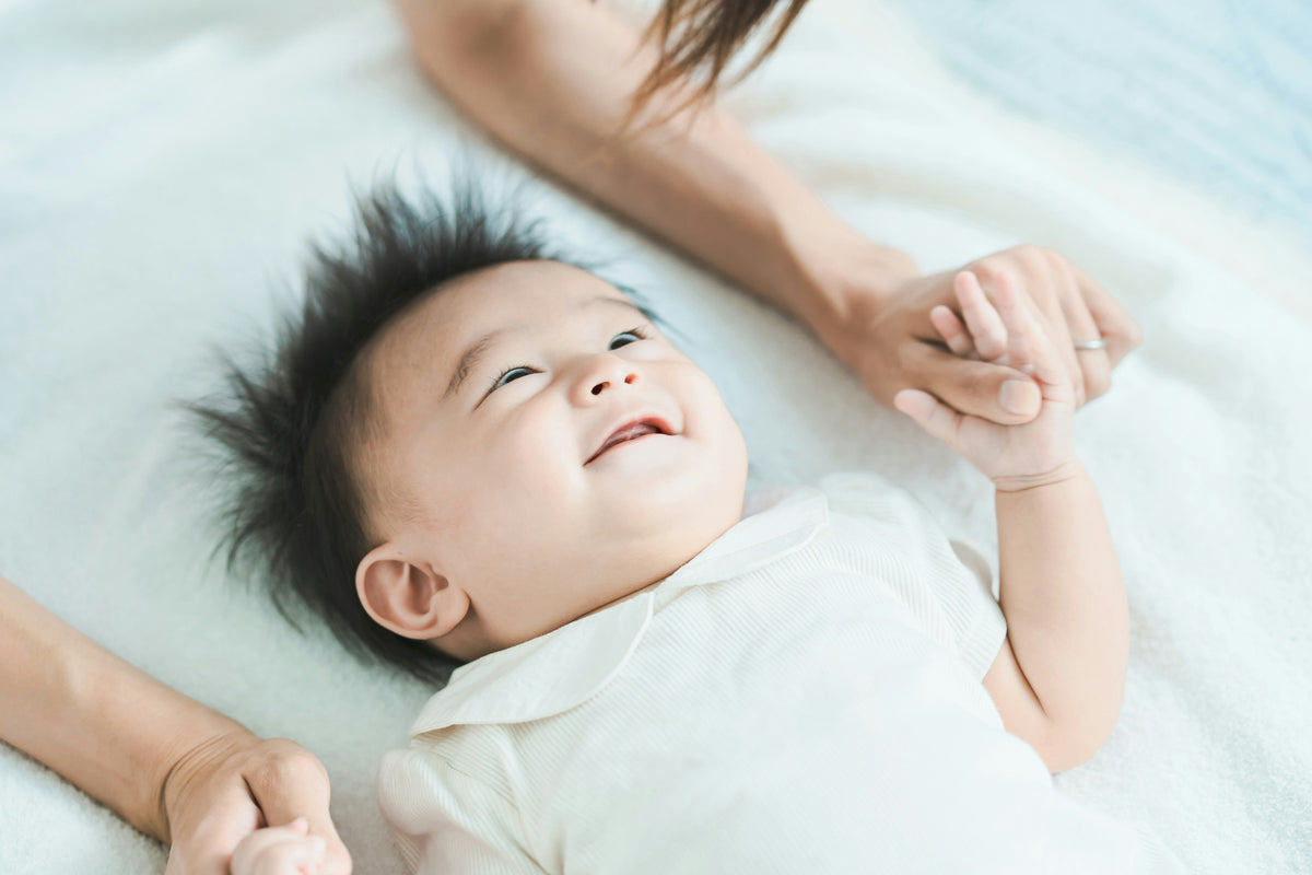 Smiling baby lying on a bed, holding mom's hand